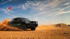 Black Toyota truck driving through a desert with a dust cloud and a vehicle safety flag visible on the car