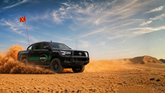 Black Toyota truck driving through a desert with a dust cloud and a vehicle safety flag visible on the car
